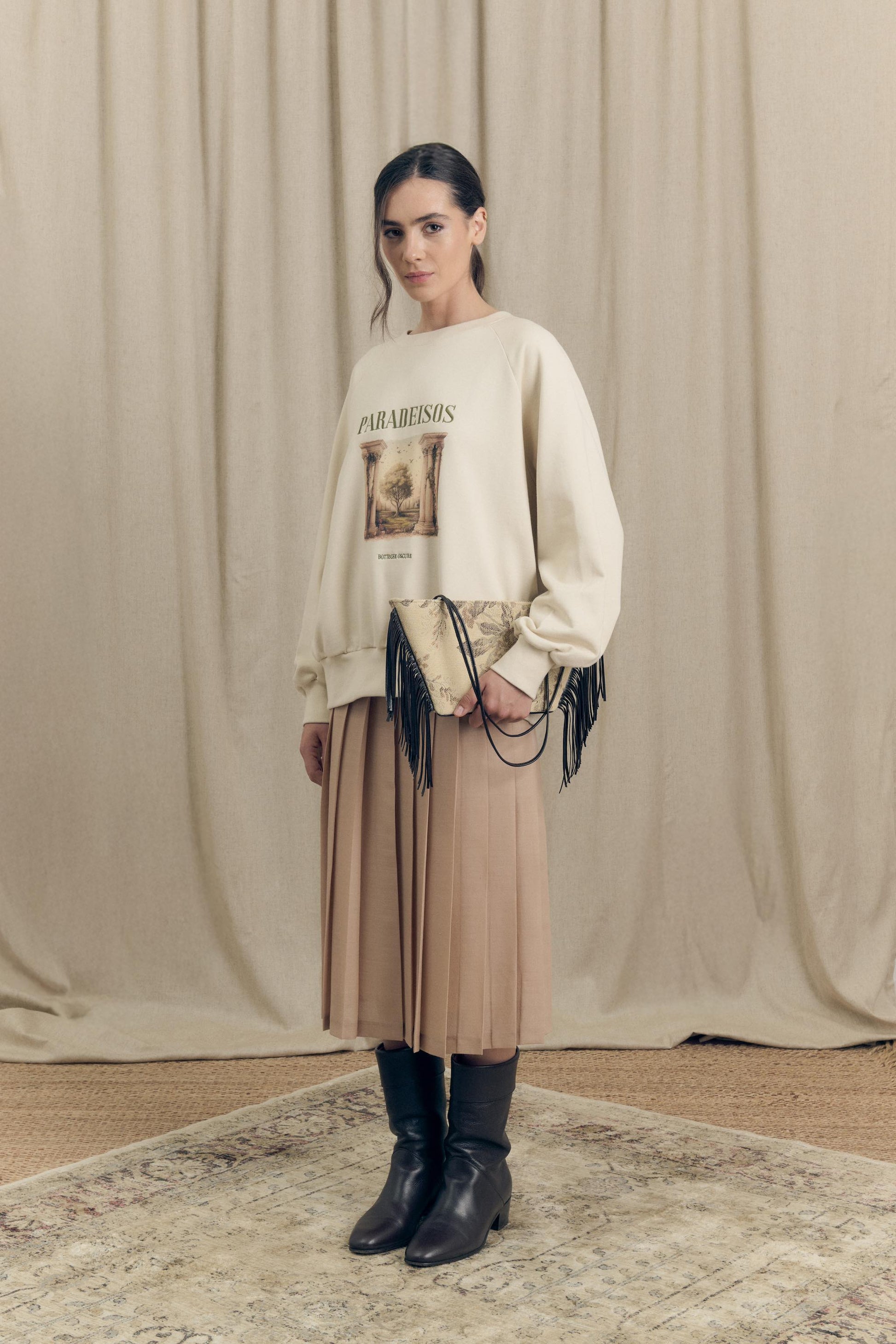 Front view of model wearing a pleated midi skirt in virgin wool paired with an oversized sweatshirt featuring Paradeisos drawing and embroidery.
