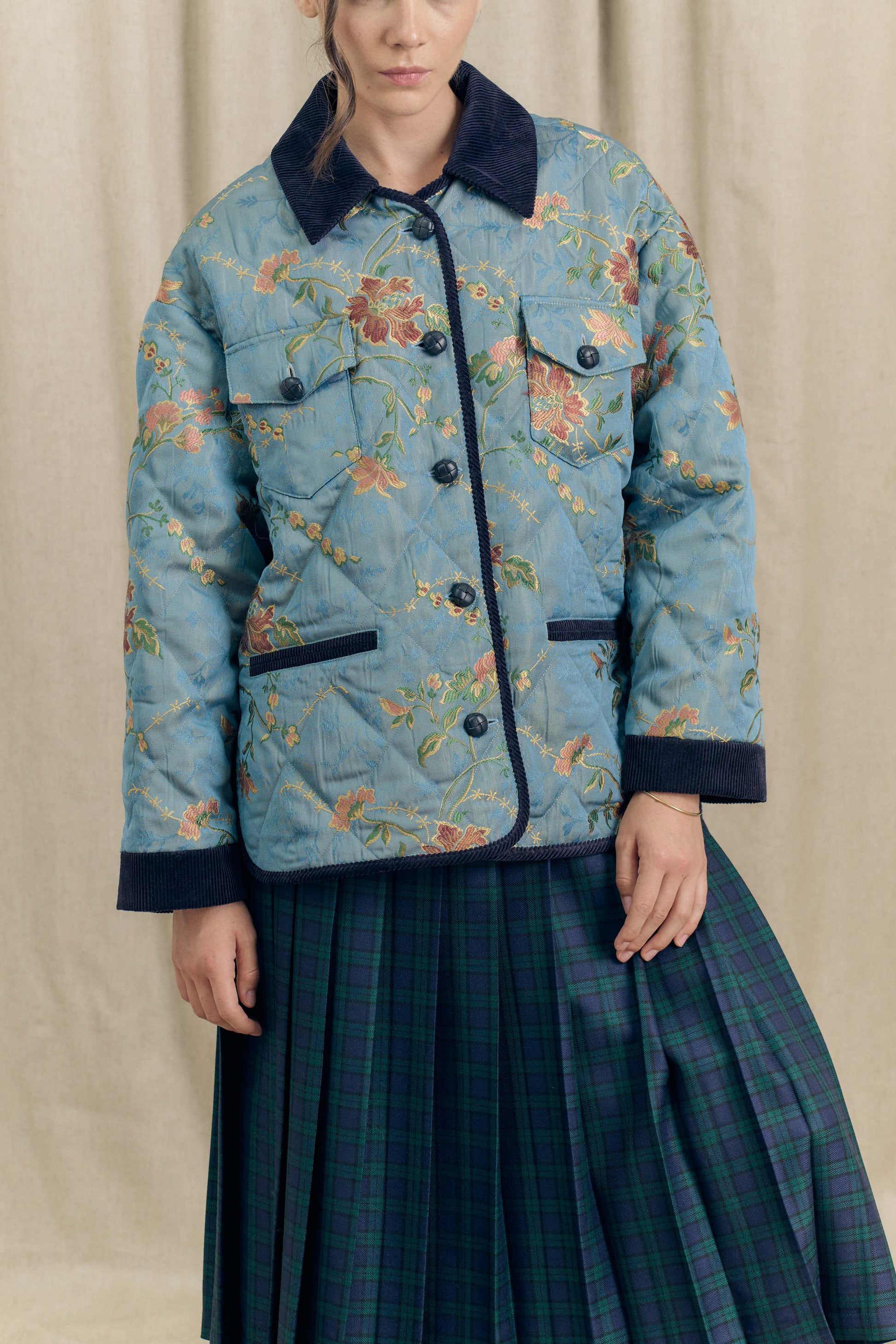 Front view of the Botteghe Oscure Look 14 floral lampas quilted jacket showing braided leather buttons and navy velvet trim, worn with a blue-green pleated skirt.