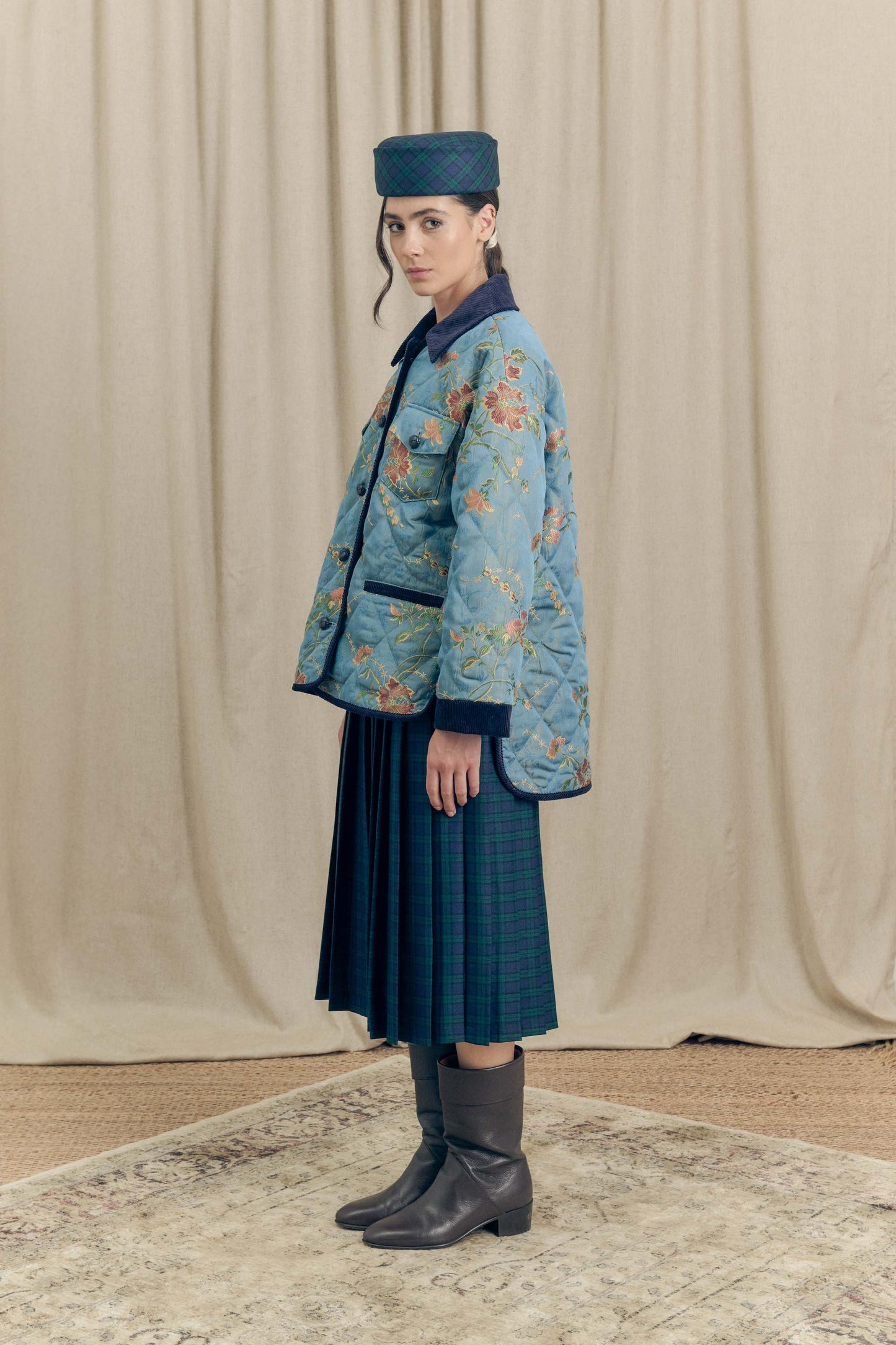 Model wearing the Botteghe Oscure Look 14 workwear quilted jacket in floral lampas cotton with navy velvet details, styled with a tartan skirt and smoking hat.