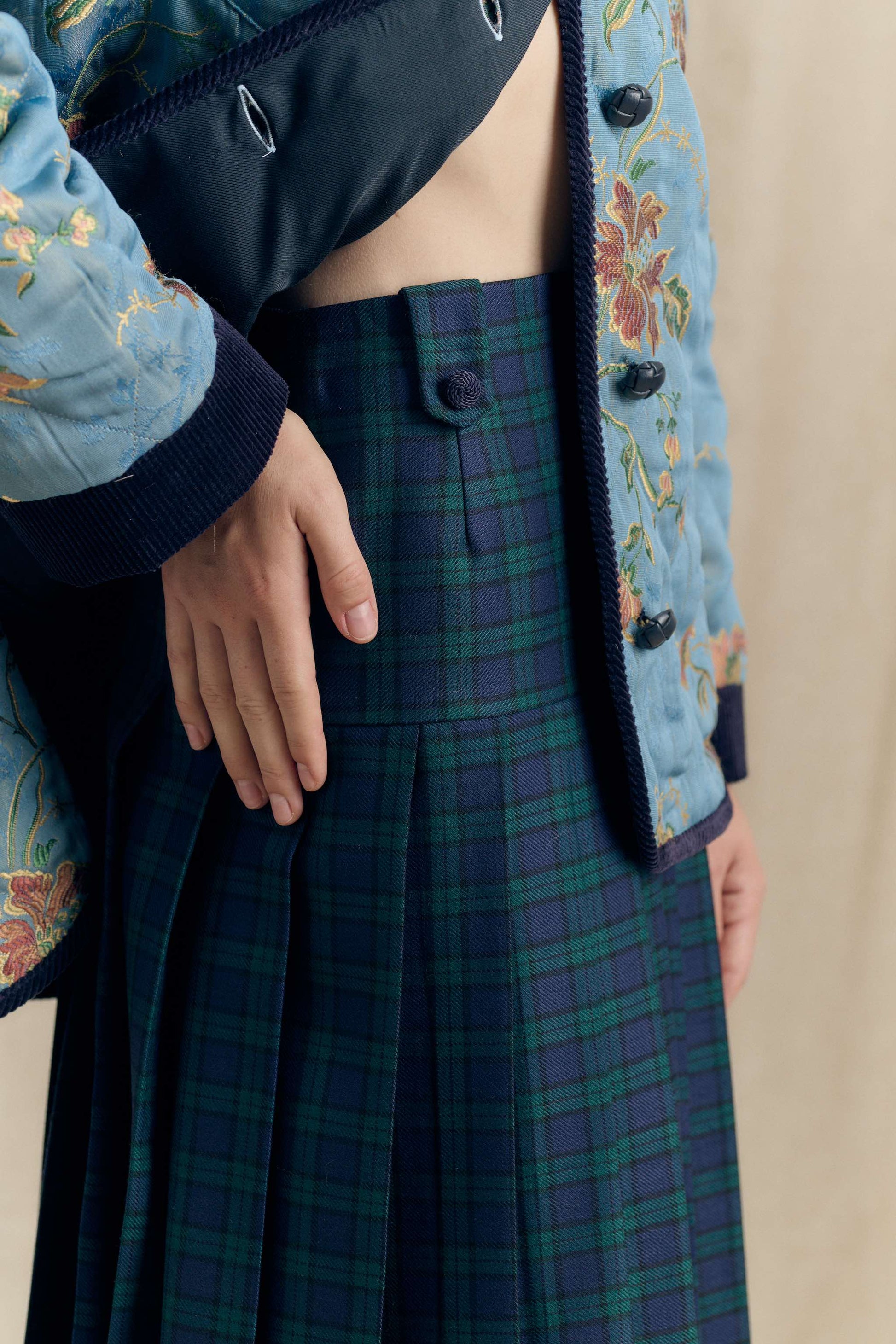 Close-up of the Botteghe Oscure Look 14 Academia skirt in Black Watch wool tartan, showing viscose passementerie button and pleated construction paired with the floral quilted jacket.
