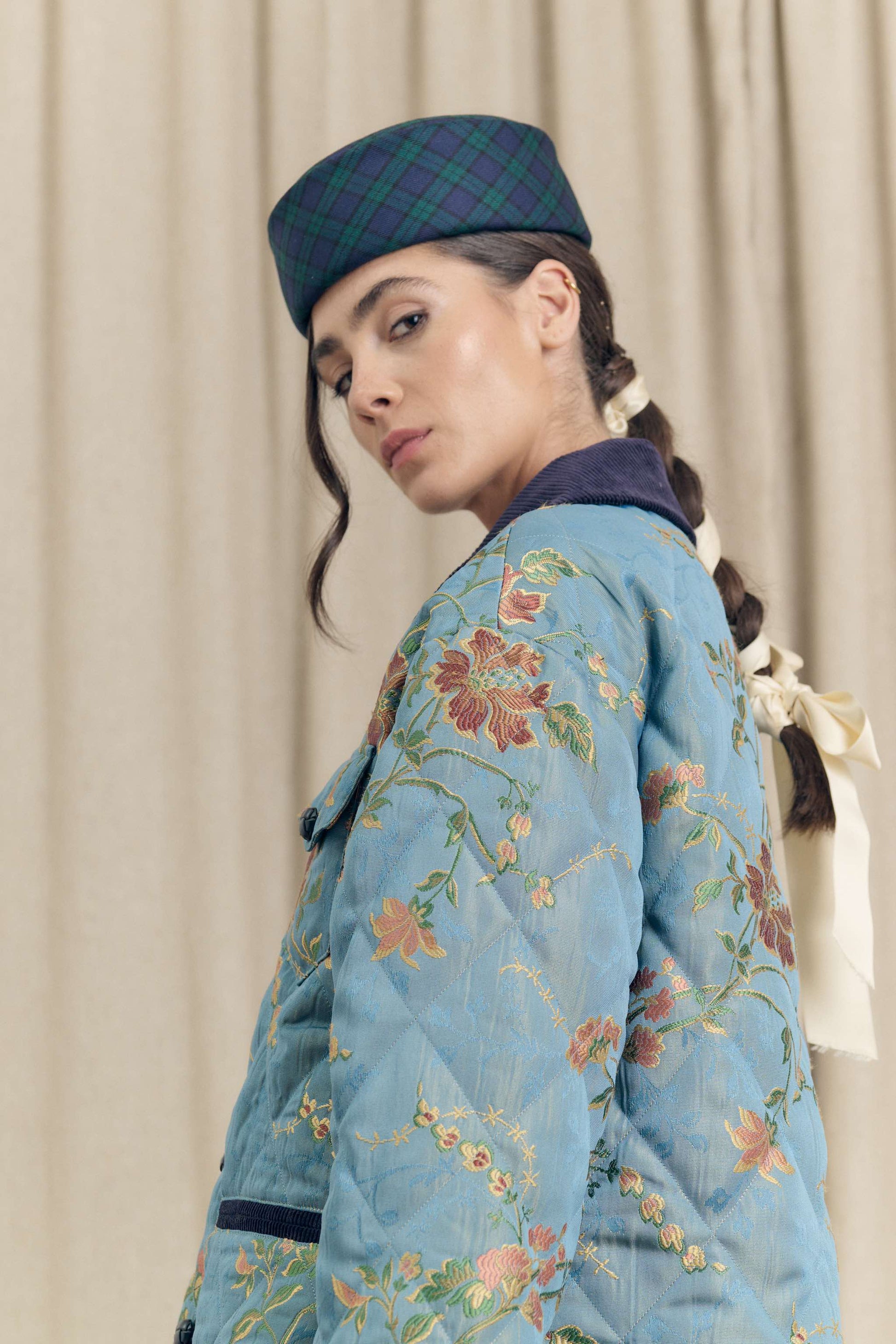 Model wearing the Botteghe Oscure handcrafted pillbox hat in Black Watch tartan wool with the floral quilted jacket in blue lampas, Look 14.