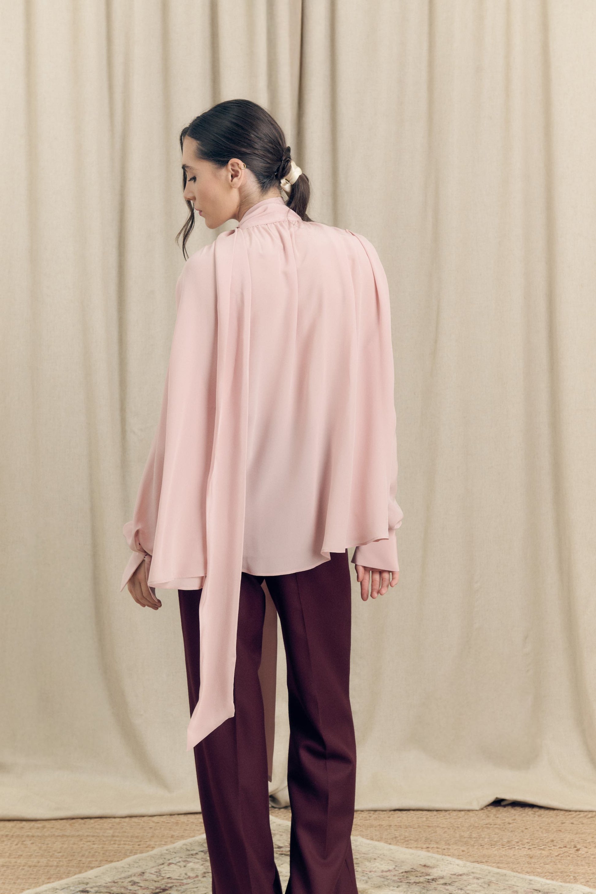 Back view of model wearing silk crepe de chine blouse with lavallière bow and ruched shoulders in blush pink by Botteghe Oscure
