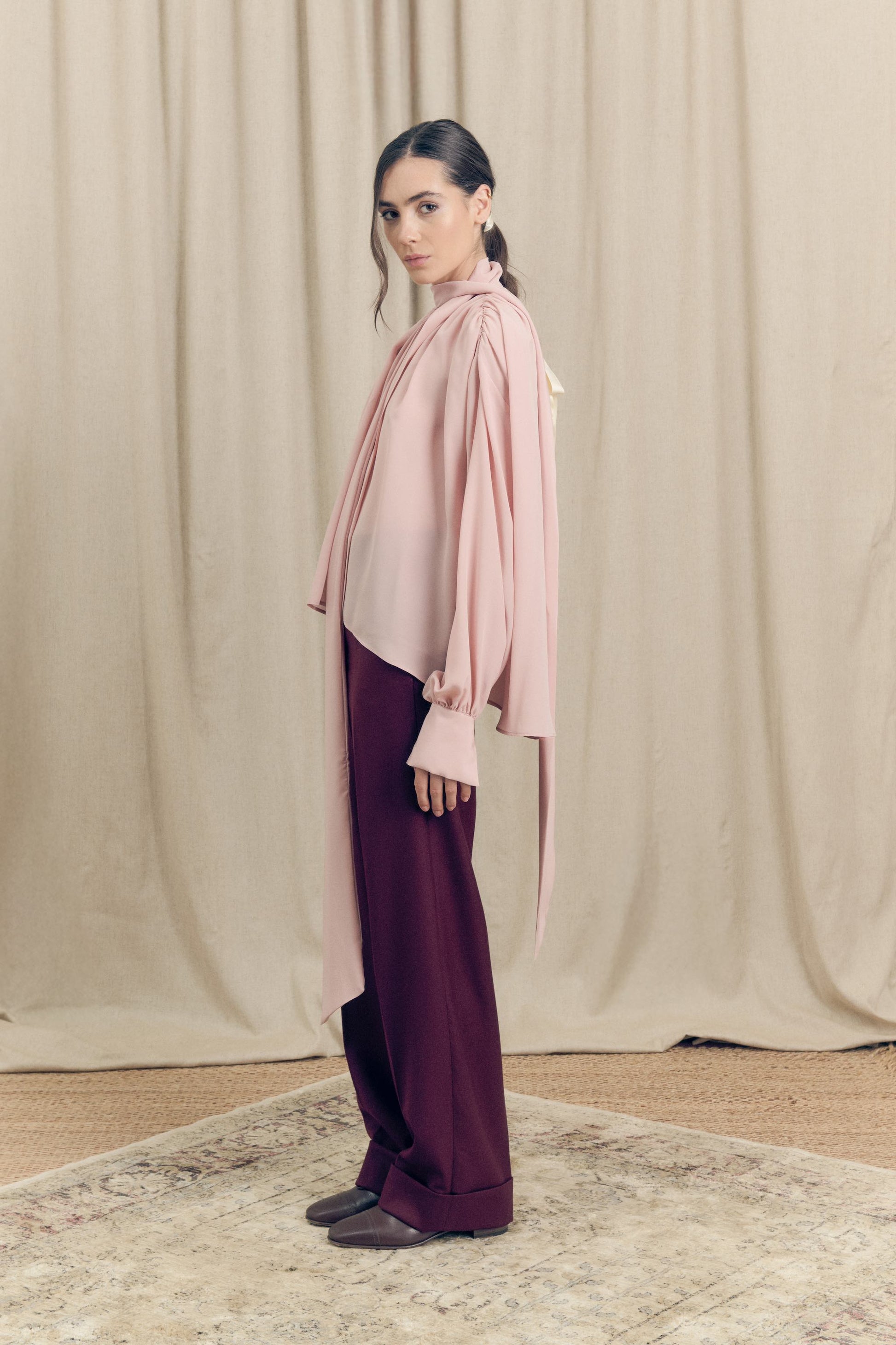 Side view of model wearing silk pink blouse and t burgundy tailored flare cut wool trousers by Botteghe Oscure