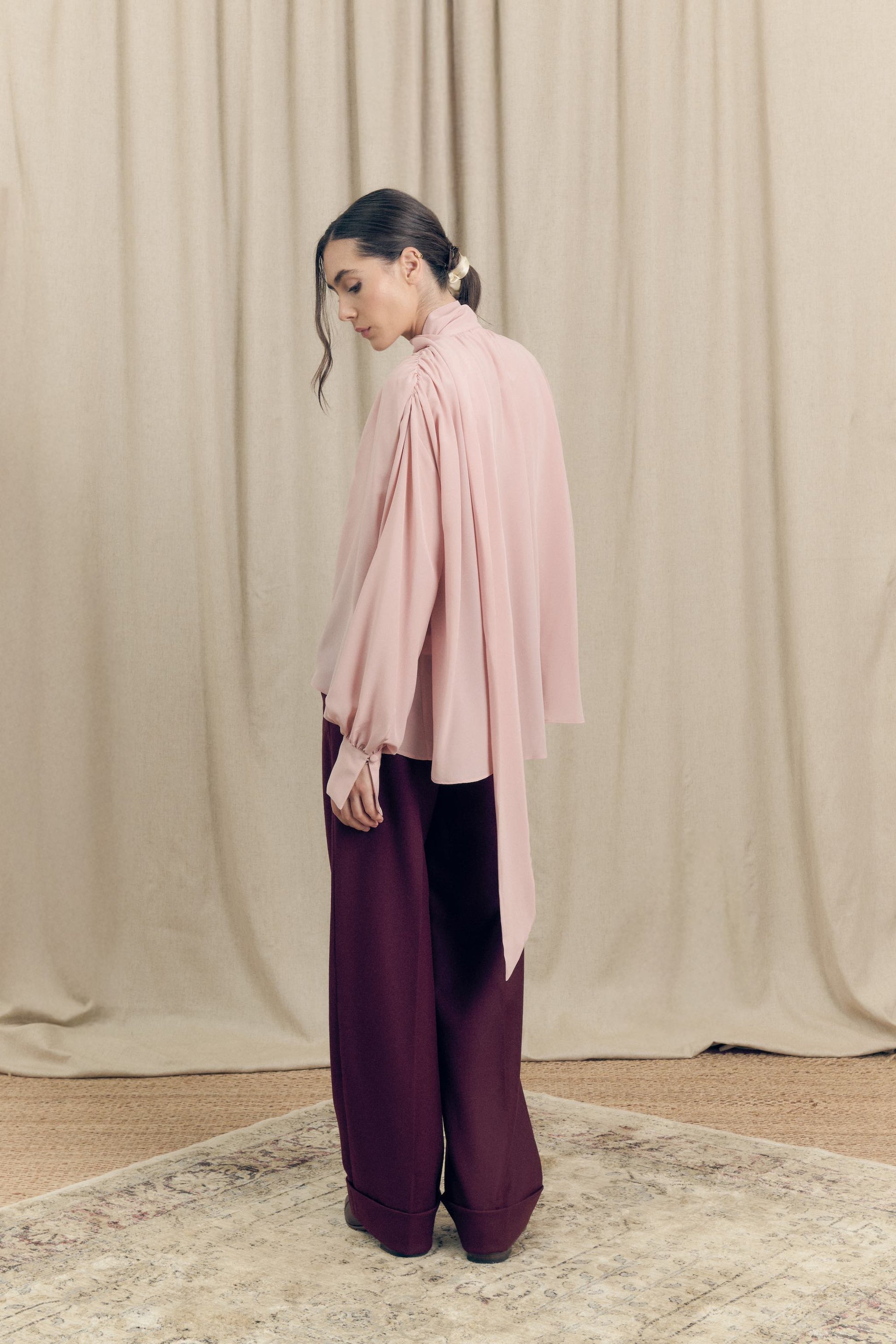 Back view of model wearing silk pink blouse and t burgundy tailored flare cut wool trousers by Botteghe Oscure